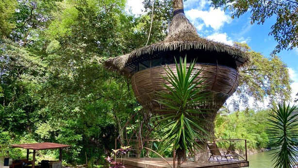 A thatched-roof structure with a wooden deck, surrounded by lush greenery and a river. - Cabanas Rio Encantado (Caldera)