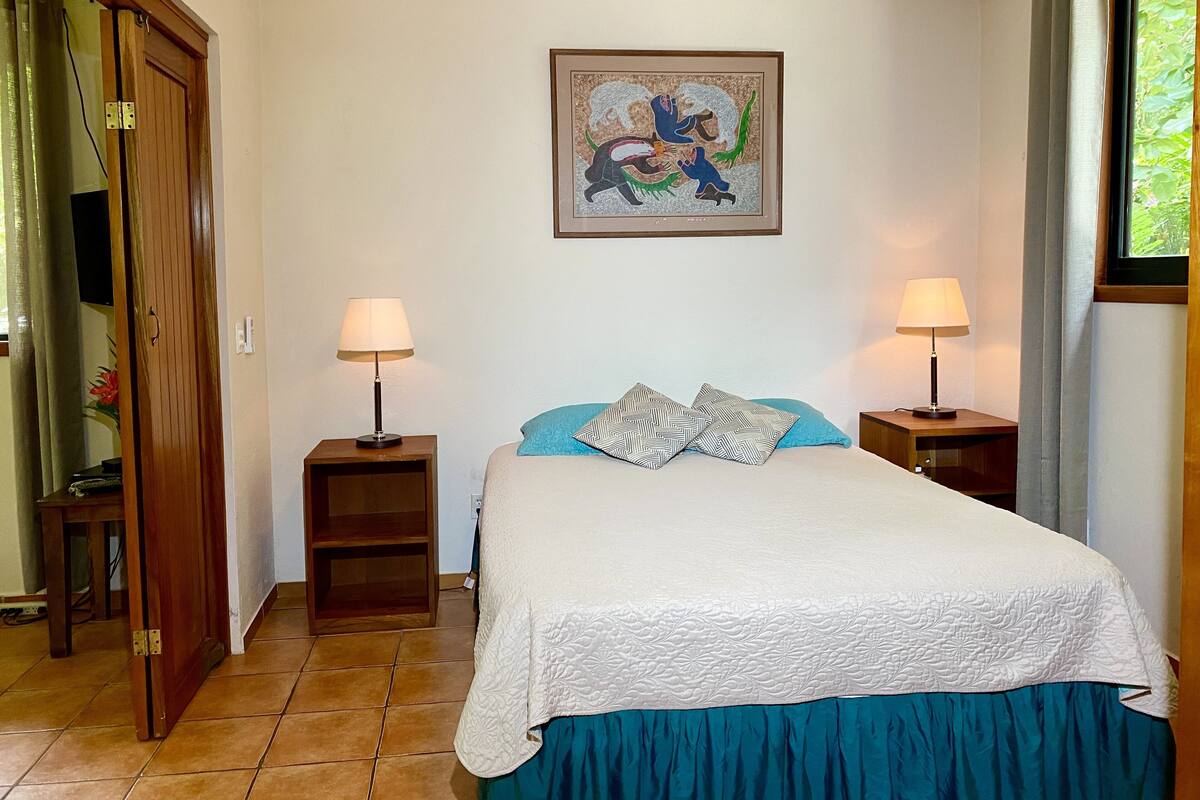 A bedroom with a bed, two bedside tables with lamps, a door, a wall-mounted painting, and a window with curtains.