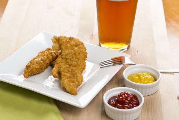Une assiette de mets panés et frits, deux petits bols de condiments et un verre d’un liquide de couleur ambrée, le tout posé sur une table en bois.