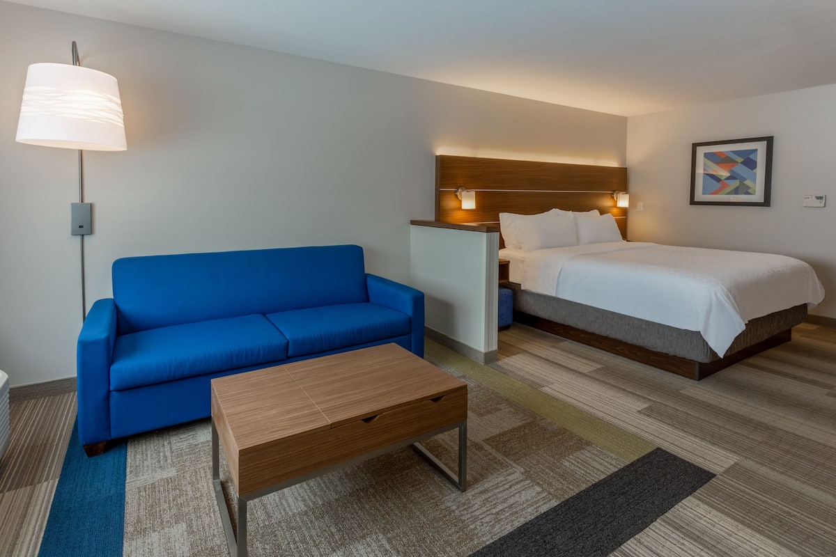 A hotel room with a blue sofa, a wooden coffee table, a bed with white bedding, a headboard with integrated lighting, and a small bedside table.