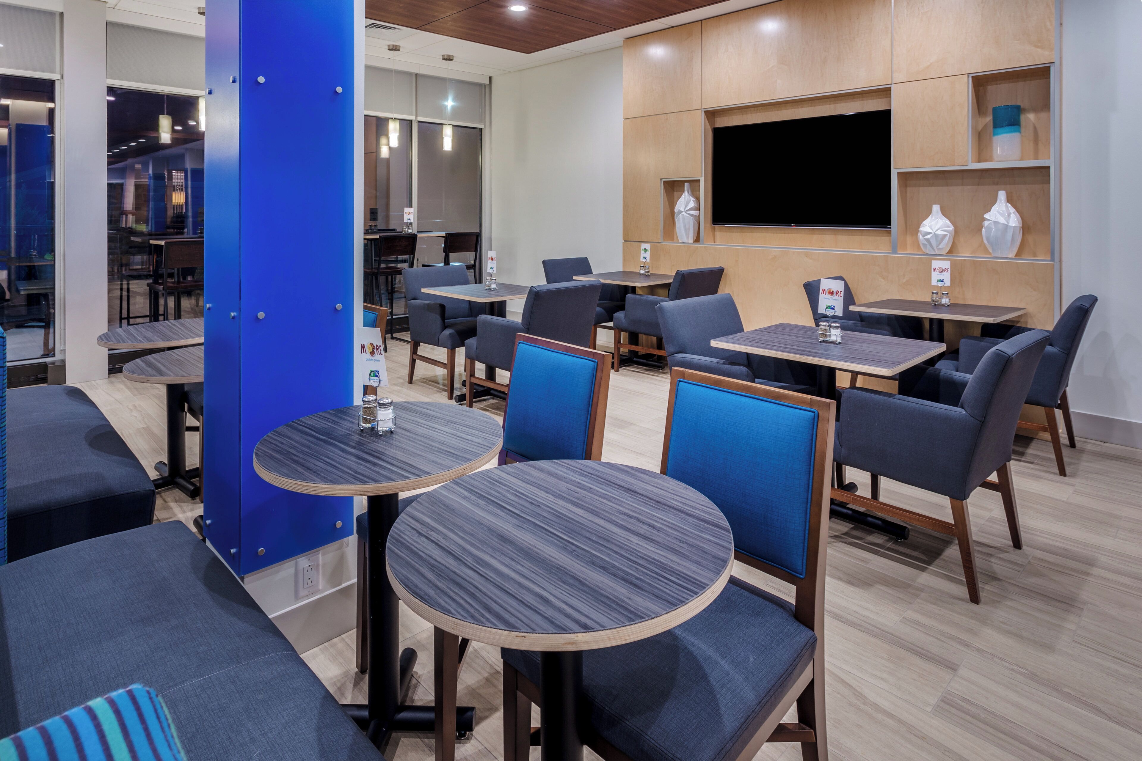 A modern restaurant with round tables and blue upholstered chairs, wooden flooring, and a flat-screen TV mounted on the wall.