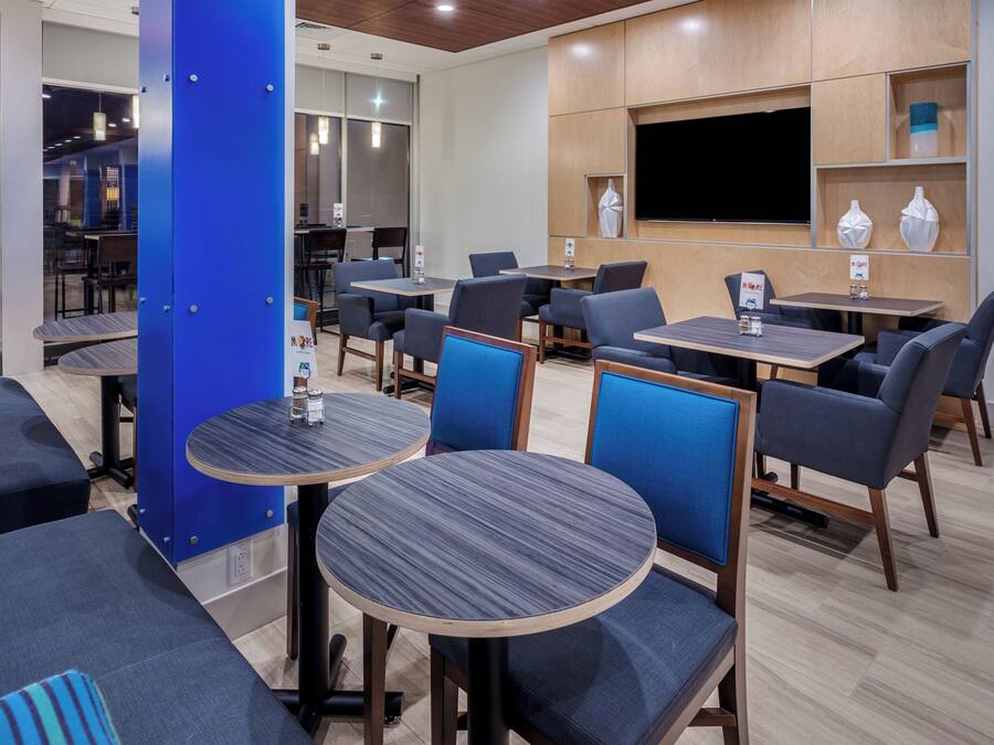 A modern restaurant with round tables and blue upholstered chairs, wooden flooring, and a flat-screen TV mounted on the wall.