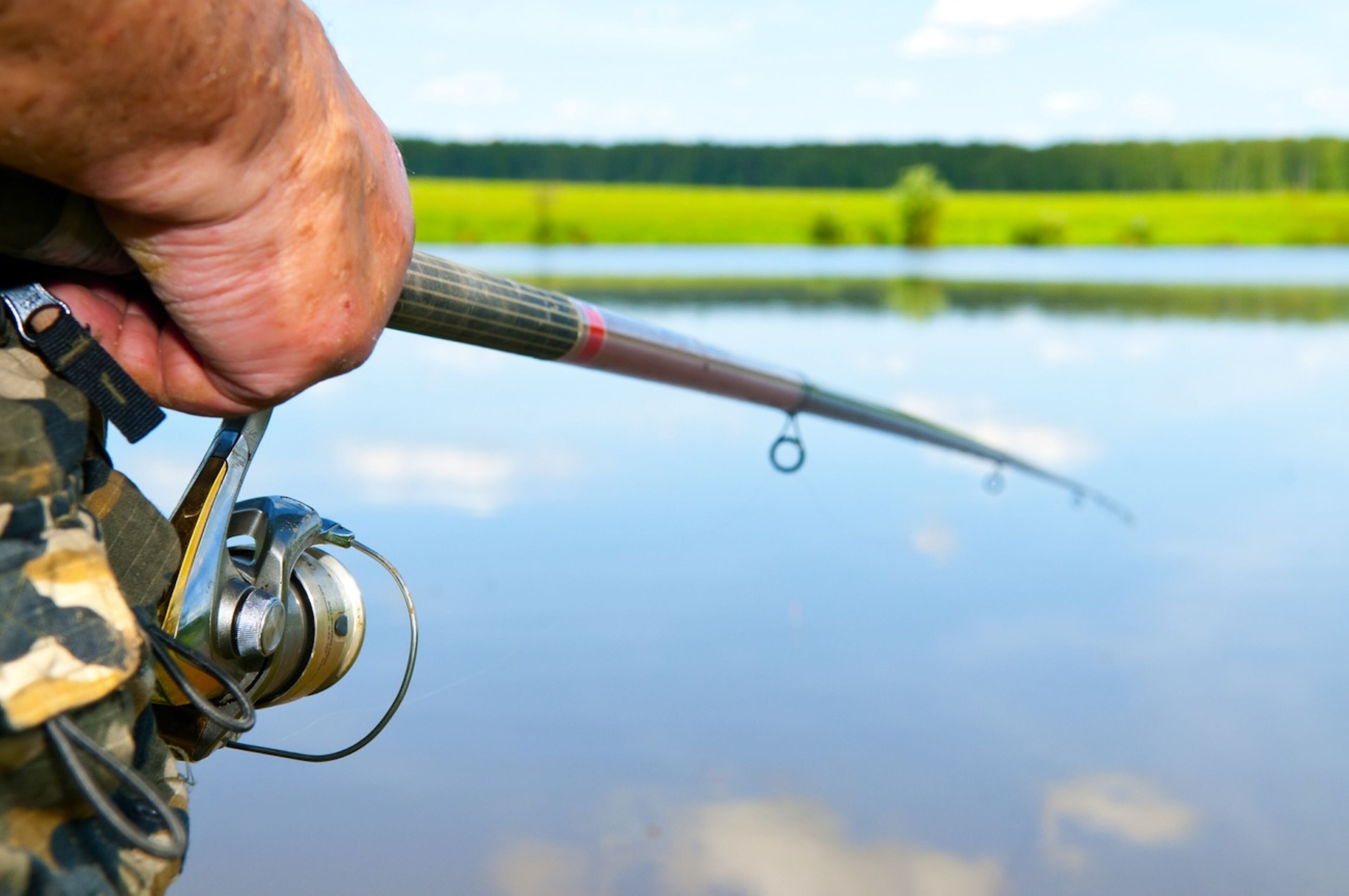 A close-up of a fishing rod with a reel, held by a person wearing camouflage clothing, with a calm lake and greenery in the background.