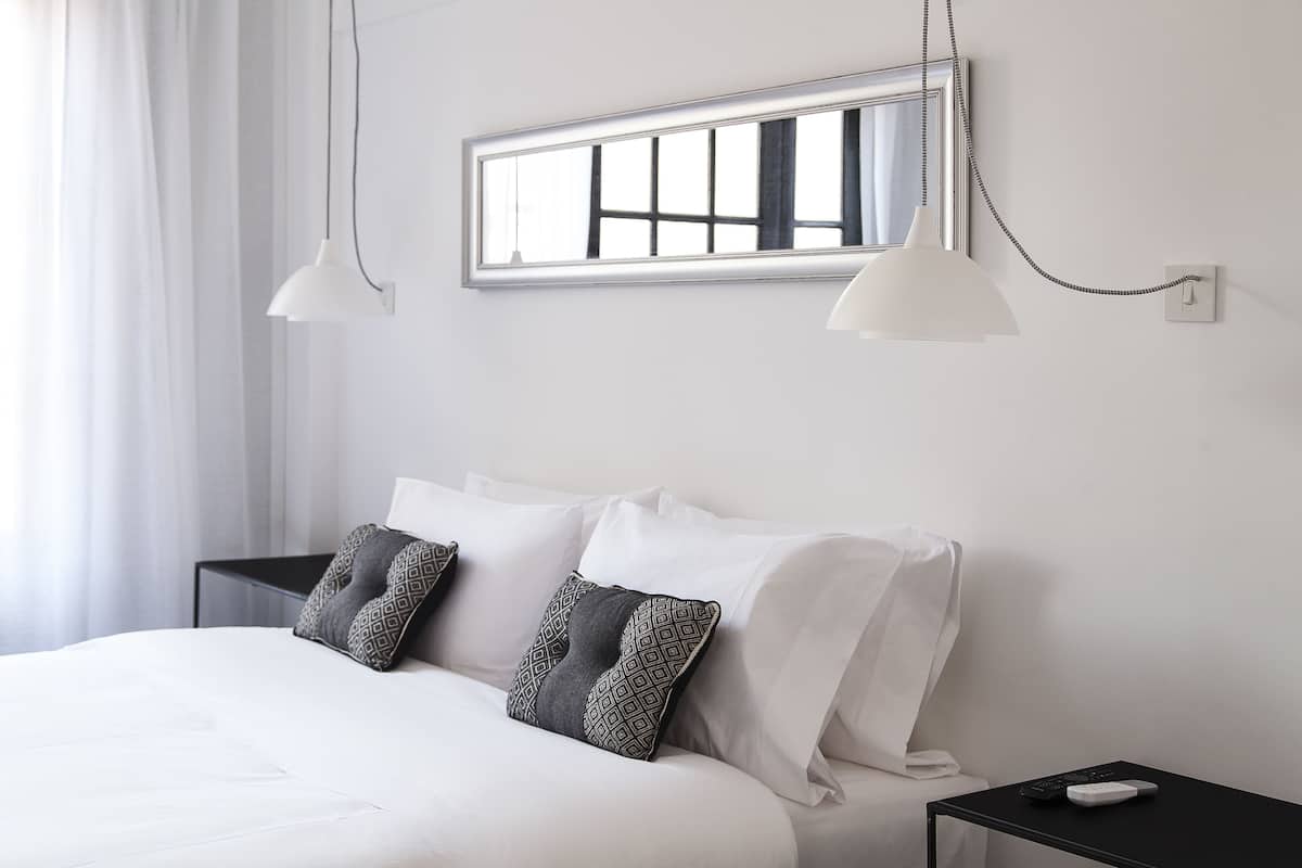 A neatly made bed with white linens and patterned pillows, a bedside table with a remote control, and a window with a view of the outside.
