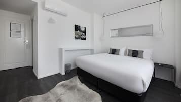 A modern bedroom with a large bed, a bedside table, a wall-mounted mirror, and a small trash bin.
