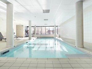 Une piscine intérieure à l’eau claire, entourée de murs et de colonnes recouverts de carreaux, avec des fenêtres laissant entrer la lumière naturelle.