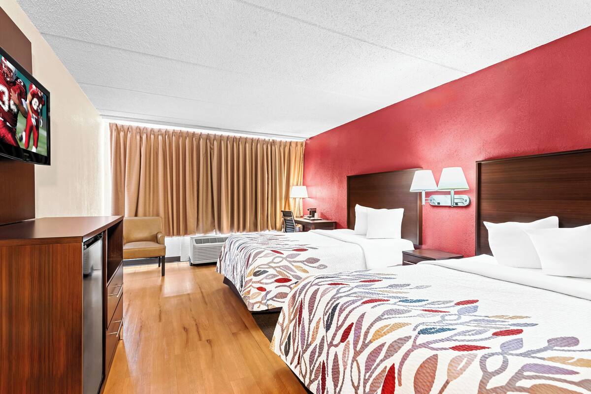 A hotel room with two beds, a desk, a TV, and a red accent wall.