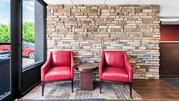 A seating area with two red chairs and a small table, a brick wall, and a view of the street through a large window. - Red Roof Inn Cortland (Cortland)