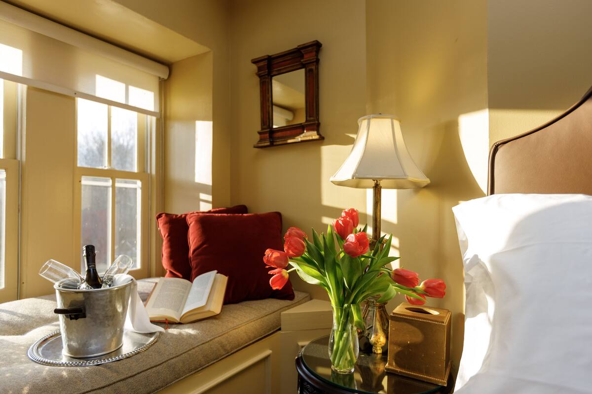 A hotel room with a bed, a bedside table with a lamp, a vase with flowers, a window, and a book on a cushioned bench.