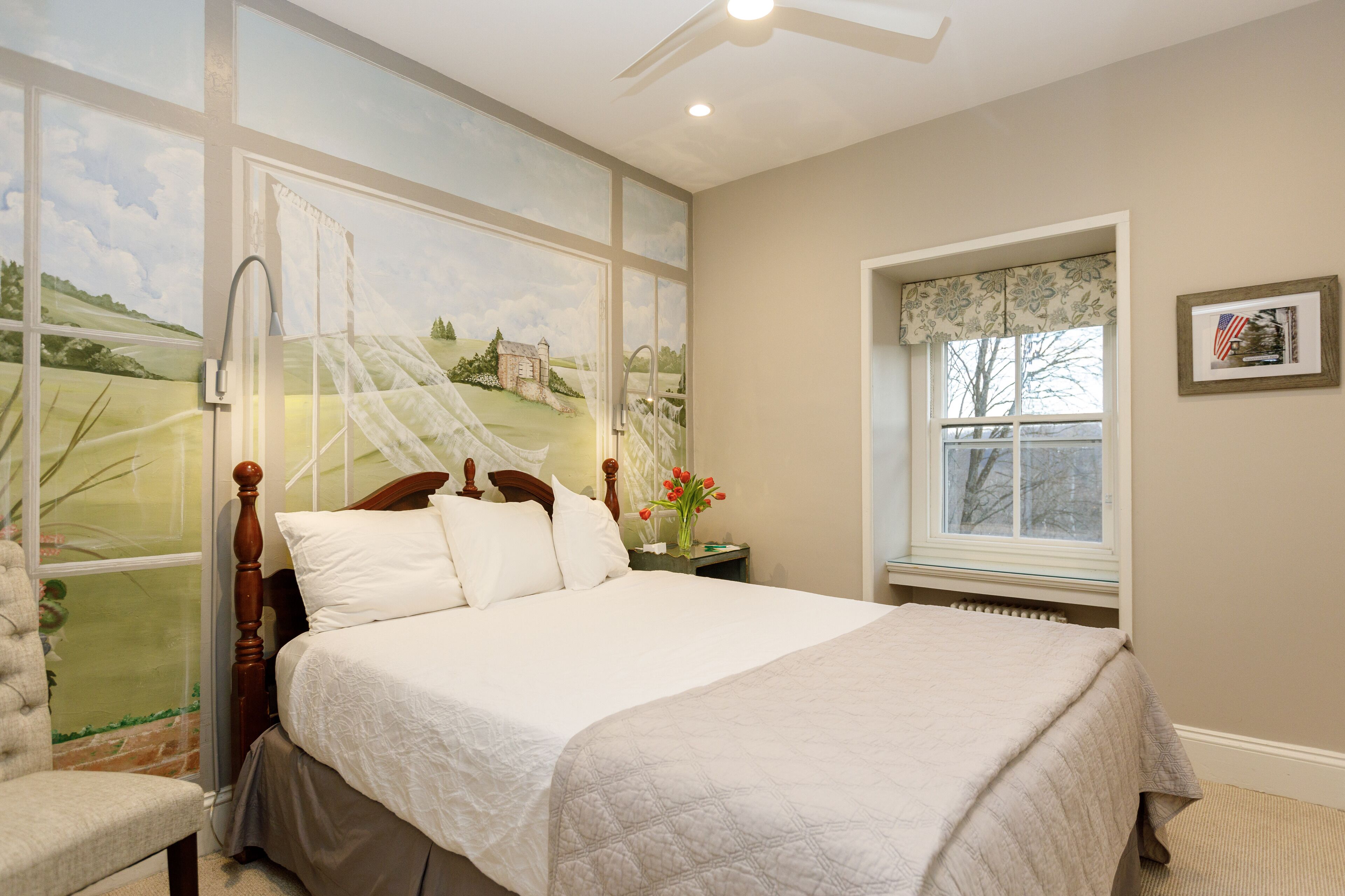 A bedroom with a large bed, a nightstand, a chair, a window with a view of a landscape, and a ceiling fan.