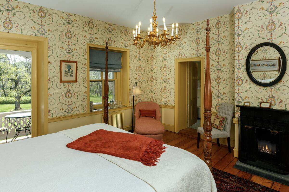 A bedroom with a large bed, a fireplace, a chandelier, and a view of the outdoors.