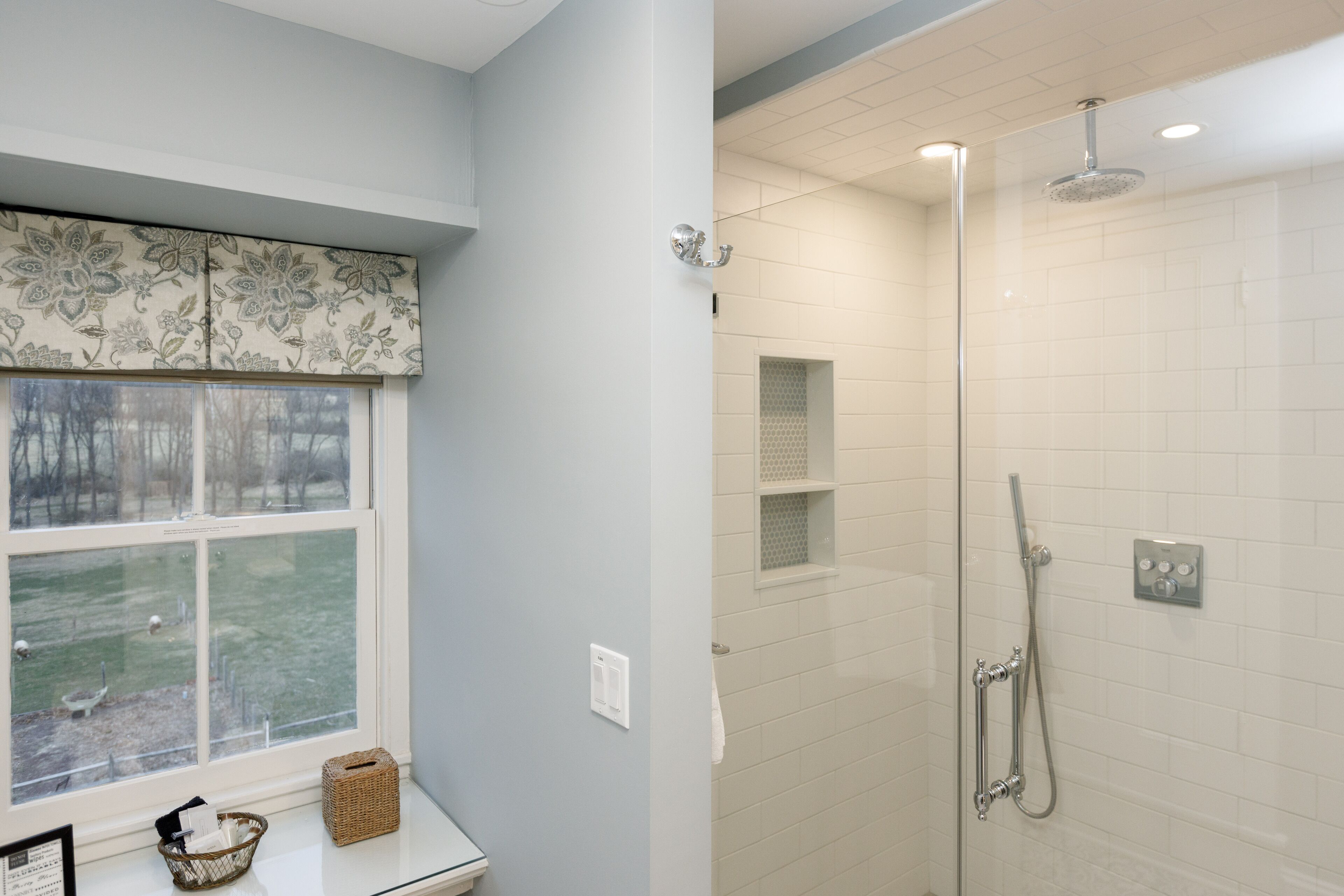 A bathroom with a shower, a window with a view of a yard, and a basket on the windowsill.