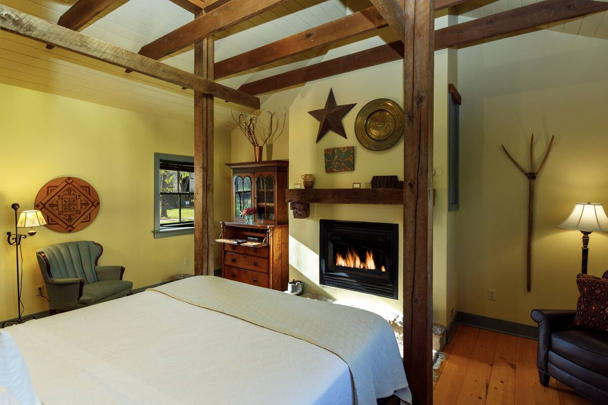 A bedroom with a large bed, wooden beams, a fireplace, and a view of the outdoors.