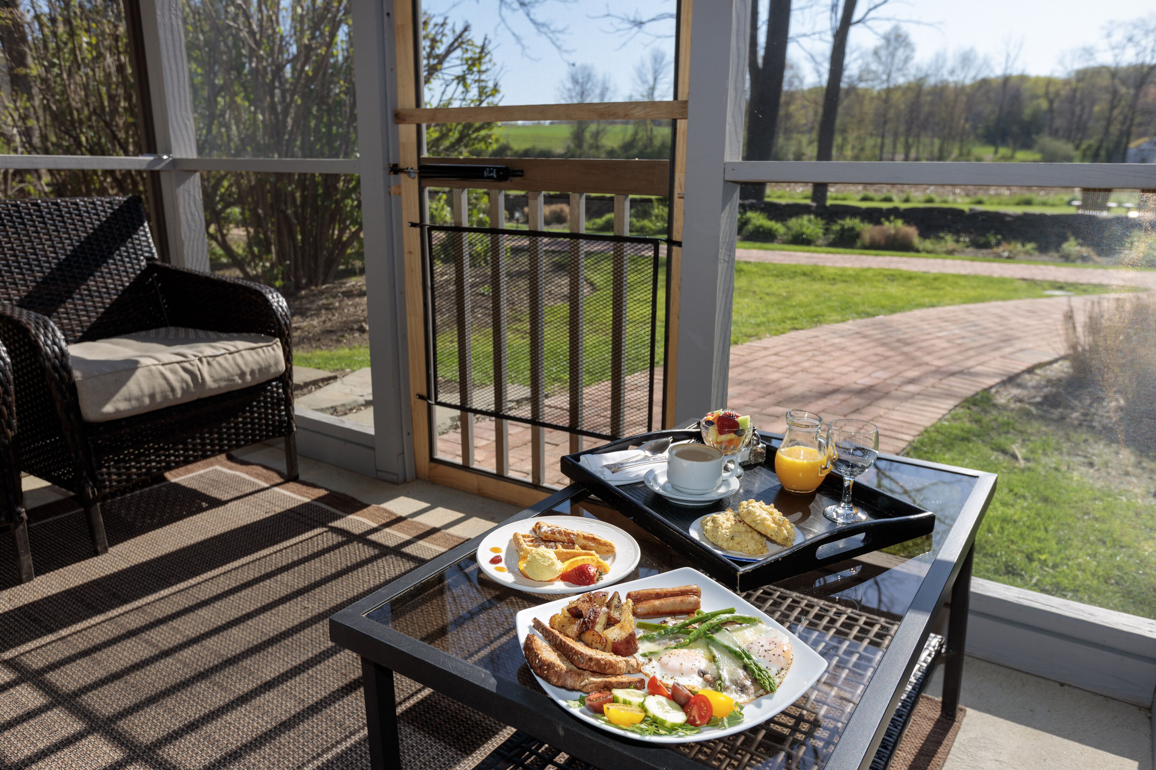 Une table de petit-déjeuner avec des assiettes remplies de nourriture, une tasse de café et un verre de jus d’orange, le tout placé sur un balcon d’où l’on peut admirer un espace verdoyant et des arbres.