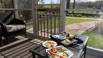 Une table de petit-déjeuner avec des assiettes remplies de nourriture, une tasse de café et un verre de jus d’orange, le tout placé sur un balcon d’où l’on peut admirer un espace verdoyant et des arbres.