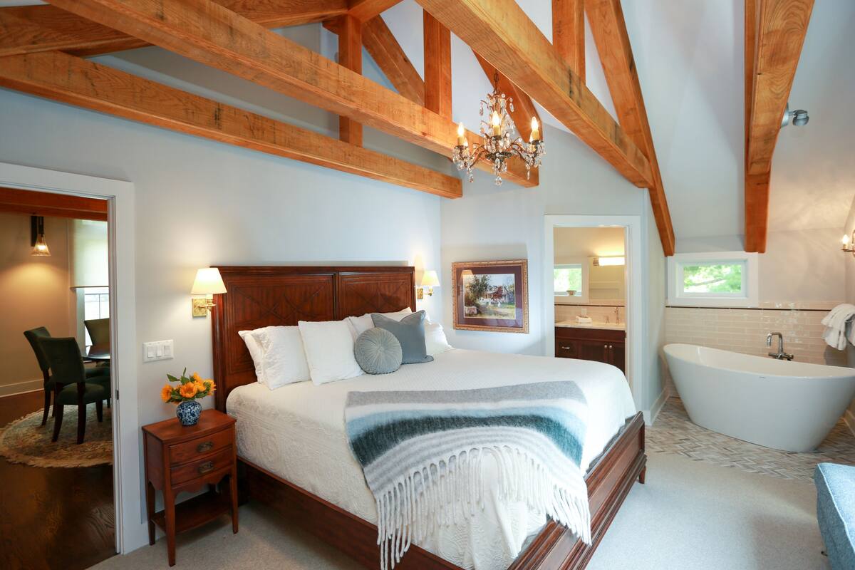 A bedroom with a large bed, a wooden ceiling, a chandelier, a nightstand, a chair, a desk, a bathroom with a bathtub, and a window.