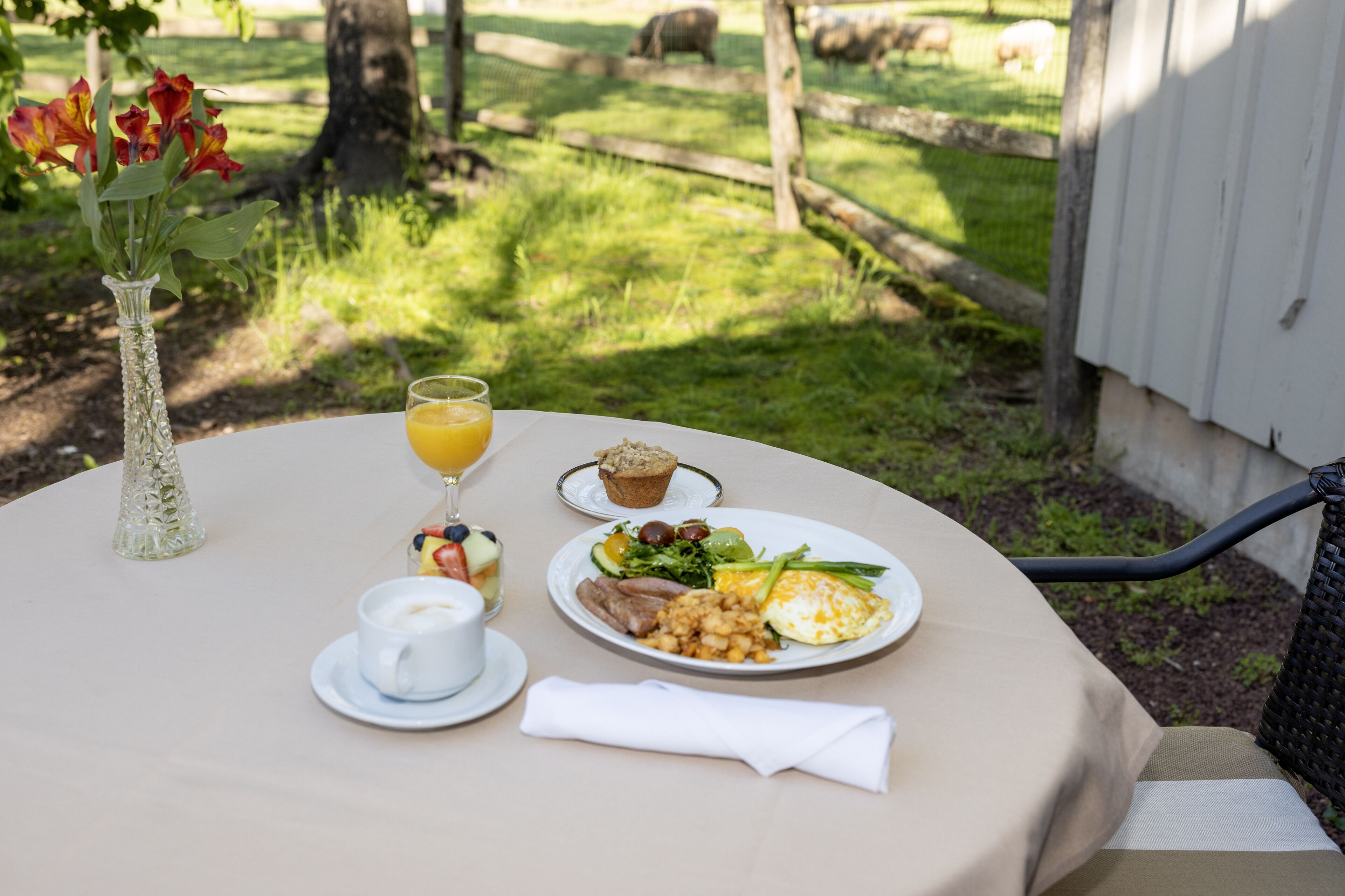 A round table with a white tablecloth, a glass of orange juice, a white cup, a plate of food, a small vase with red flowers, and a napkin.