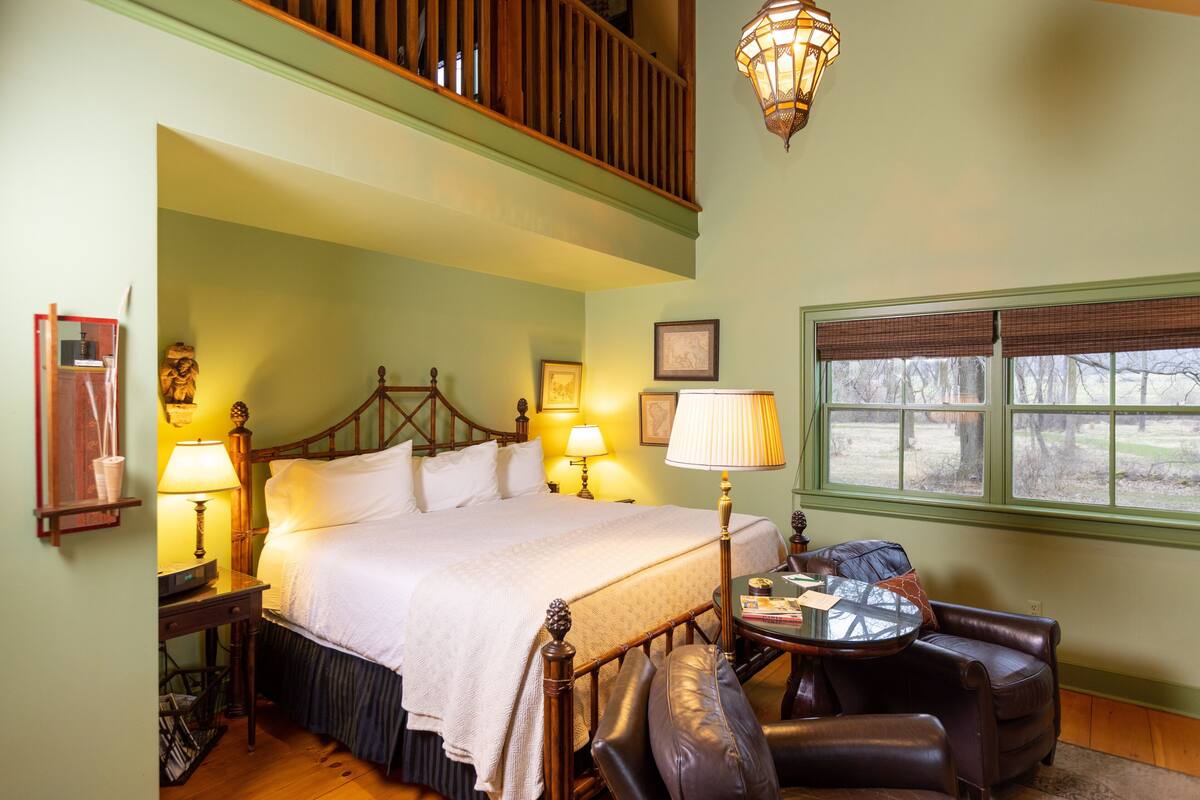 A bedroom with a large bed, a wooden headboard, a nightstand with a lamp, a leather armchair, a small round table, and a window with blinds.