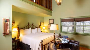 A bedroom with a large bed, a wooden headboard, a nightstand with a lamp, a leather armchair, a small round table, and a window with blinds.