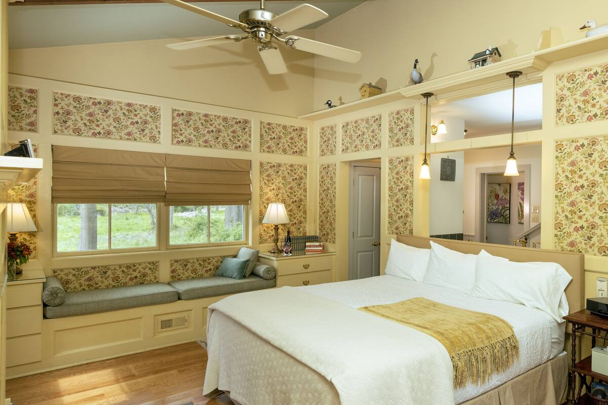 A bedroom with a large bed, a window seat, a ceiling fan, and floral wallpaper.