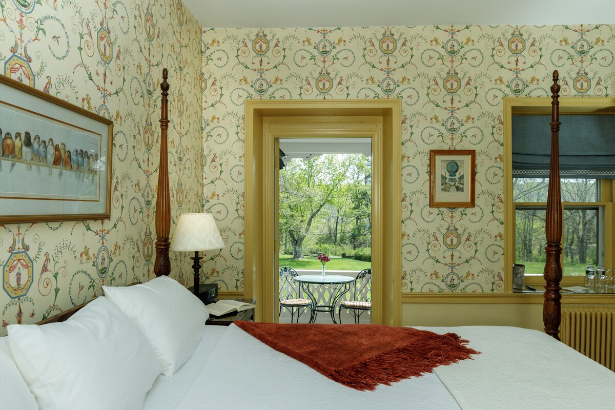Una habitación de hotel con una cama, una mesita de noche, una lámpara, un cuadro en la pared y una ventana con vistas a un jardín.