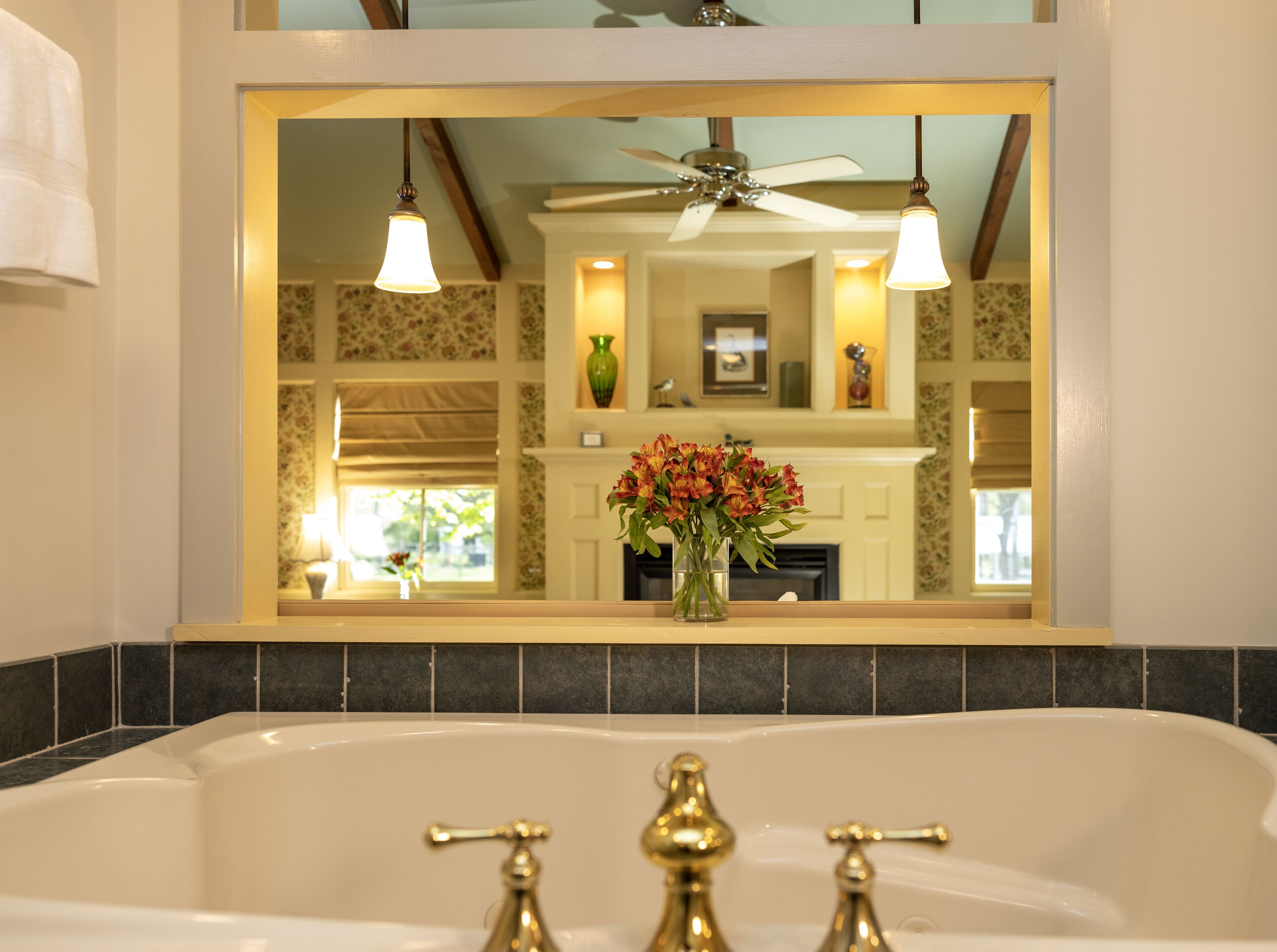 A bathroom with a bathtub, a mirror reflecting a room with a fireplace, and a ceiling fan.