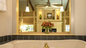 A bathroom with a bathtub, a mirror reflecting a room with a fireplace, and a ceiling fan.