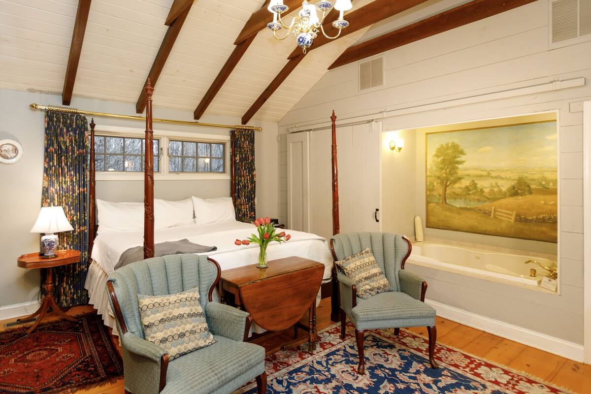 A bedroom with a four-poster bed, a large window, a painting, and two upholstered chairs.