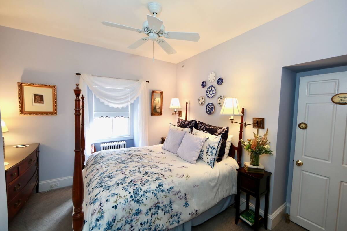 A bedroom with a four-poster bed, a ceiling fan, a white door, and a wall with decorative plates.