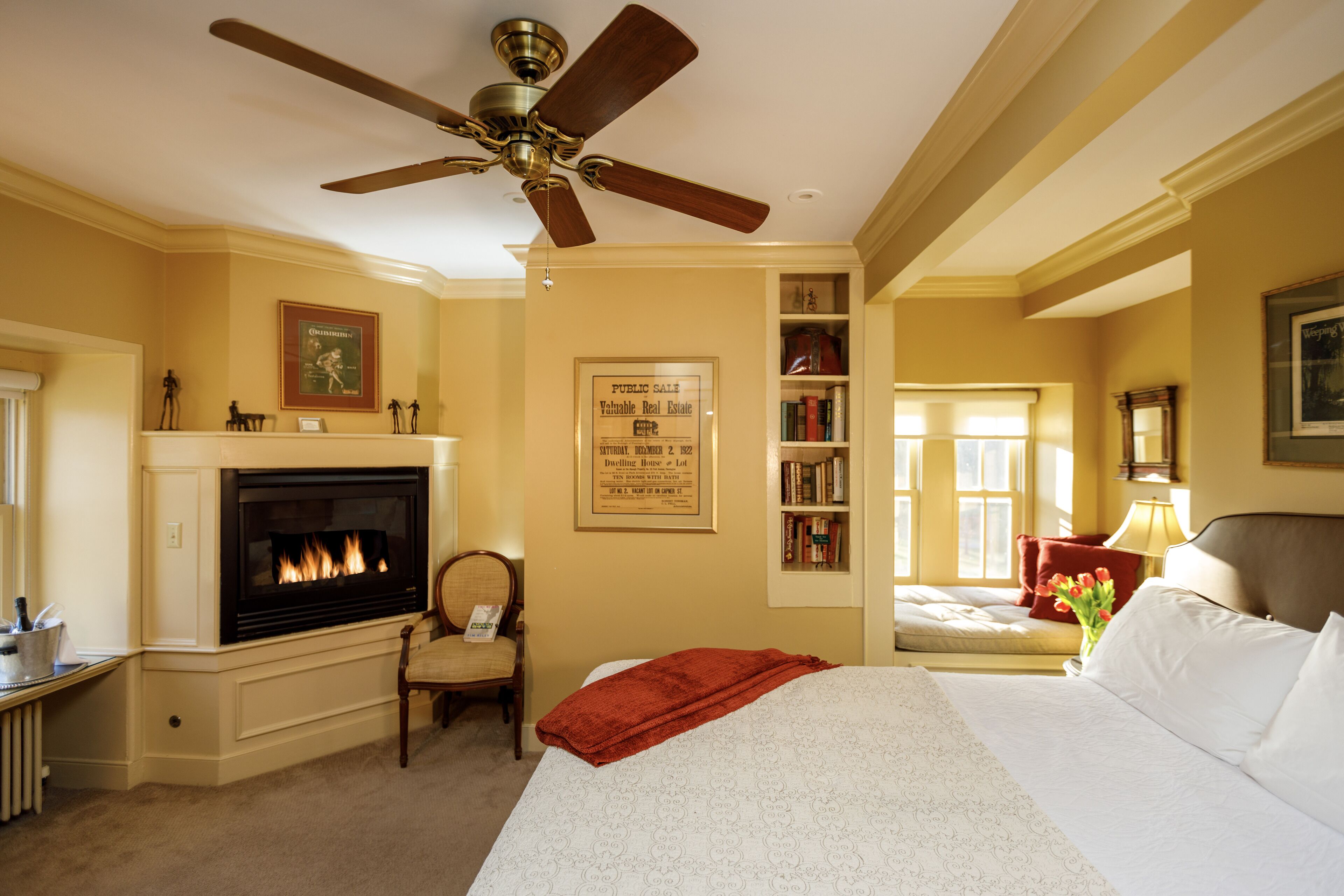 A hotel room with a fireplace, a bed, a bookshelf, and a ceiling fan.