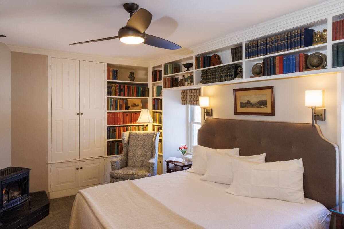 A bedroom with a large bed, a bookshelf, a chair, and a ceiling fan.