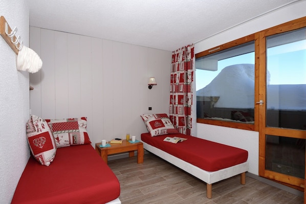 Une chambre avec deux lits simples, une petite table, une lampe fixée au mur et une fenêtre donnant sur des collines enneigées.