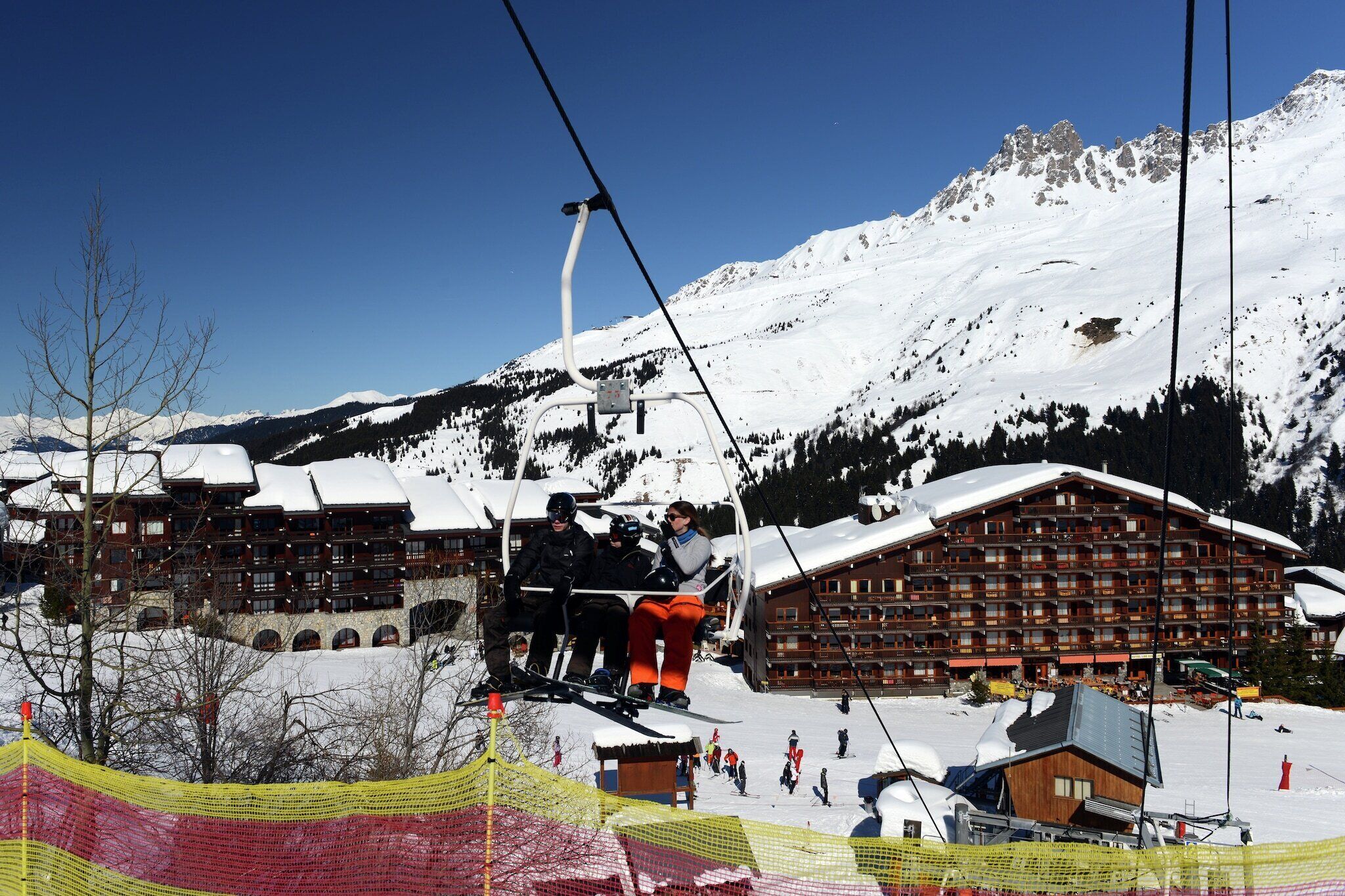 A ski lift with skiers, wooden buildings, and snow-covered mountains in the background.
