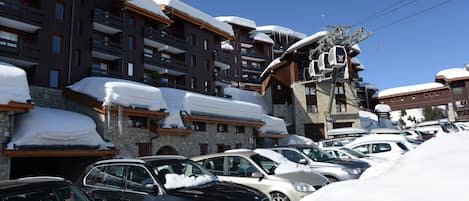Un estacionamiento cubierto de nieve con coches y un edificio con balcones y un telesilla.