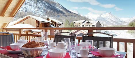 Mesa preparada para el desayuno con vista a la montaña, incluyendo cabañas de madera y picos nevados.