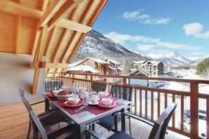 Eine Holzdiele mit einem für das Frühstück gedeckten Tisch, mit Blick auf eine Berglandschaft mit Chalets.