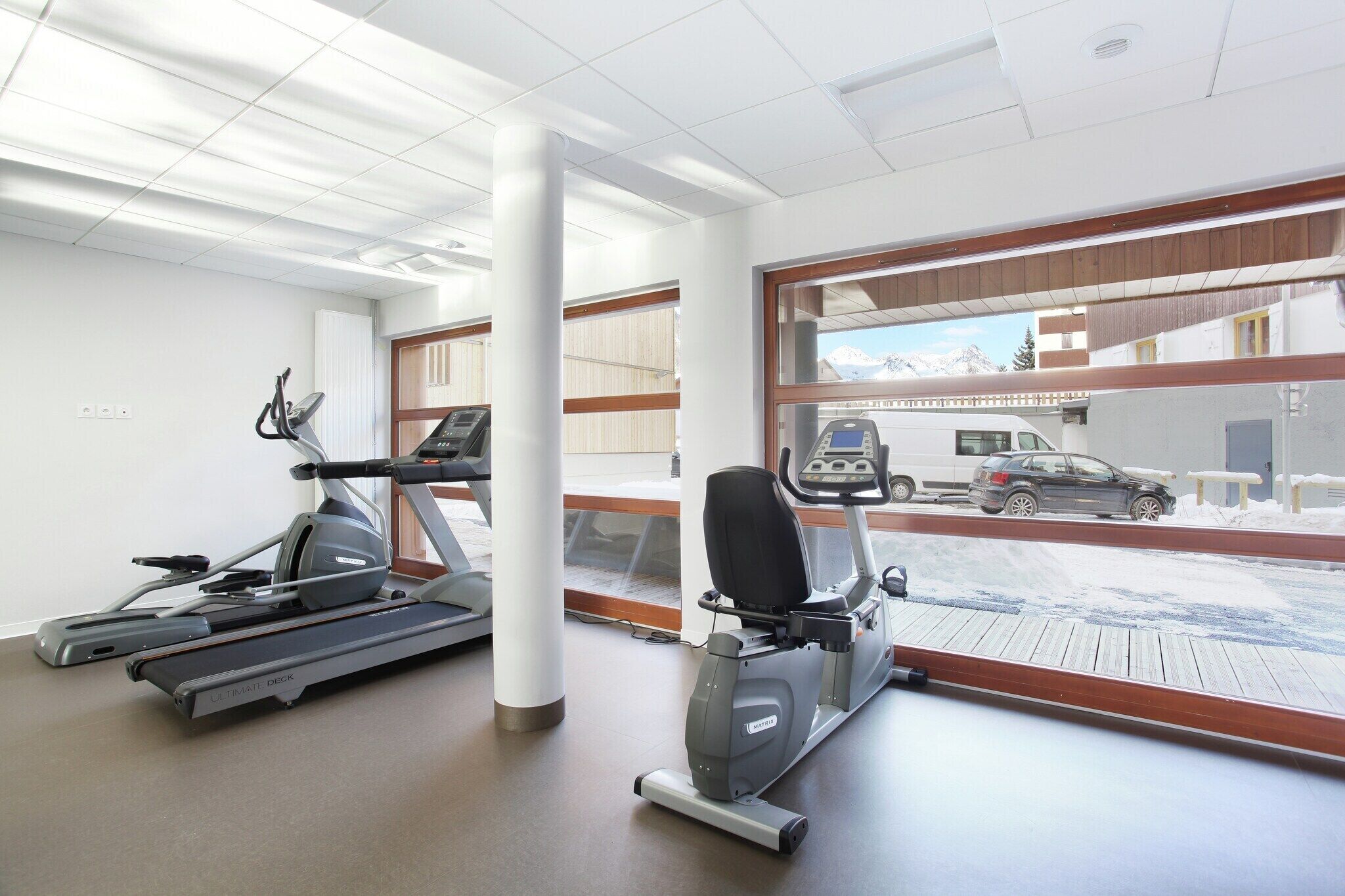 A gym with treadmills and an elliptical machine, large windows with a view of a snowy landscape and parked cars.