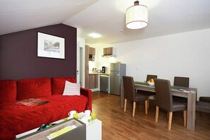 Ein kompakter Wohnraum mit einem roten Sofa, einem Esstisch mit Stühlen, einer Küchenzeile mit Kühlschrank und einer Pendelleuchte.