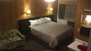 Habitación de hotel con cama, dos lámparas fijadas a la pared, una mesita de noche, una silla con estampado floral, un baño con lavamanos rojo y paneles de madera en las paredes.