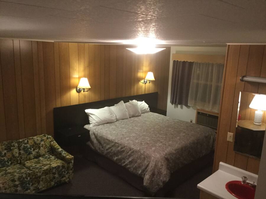 Quarto de hotel com uma cama, duas luminárias fixadas na parede, uma mesa de cabeceira, uma cadeira com padrão floral, um banheiro com pia vermelha e painéis de madeira nas paredes.