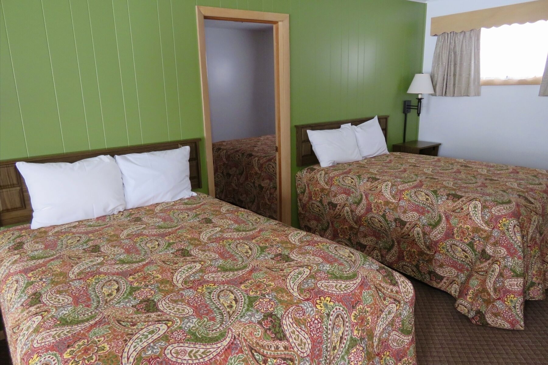 A room with two beds, each with a patterned bedspread and white pillows. There is a door leading to another room, a nightstand with a lamp, and a window with curtains.