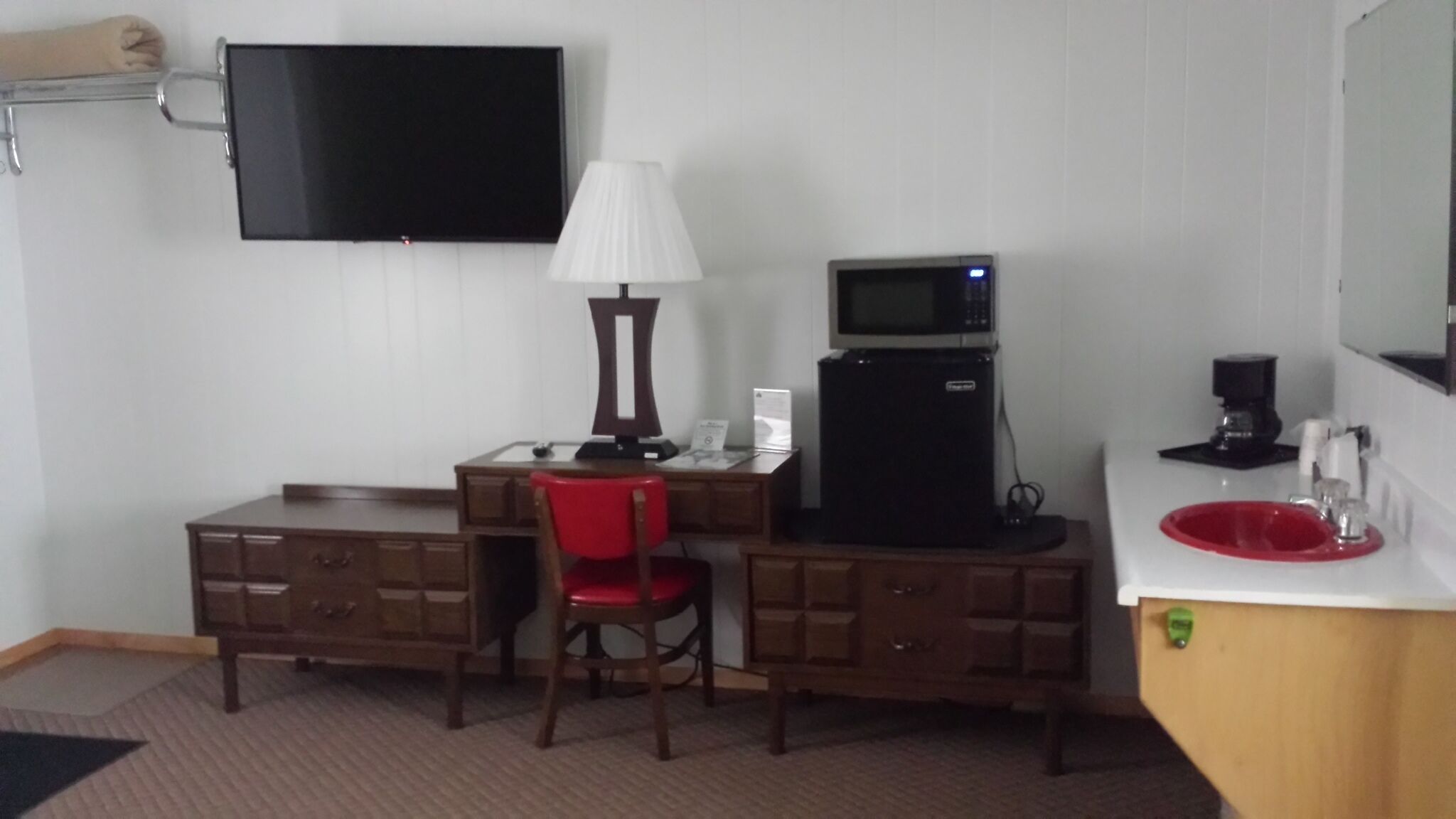A hotel room with a desk, a chair, a television, a lamp, a refrigerator, and a sink.