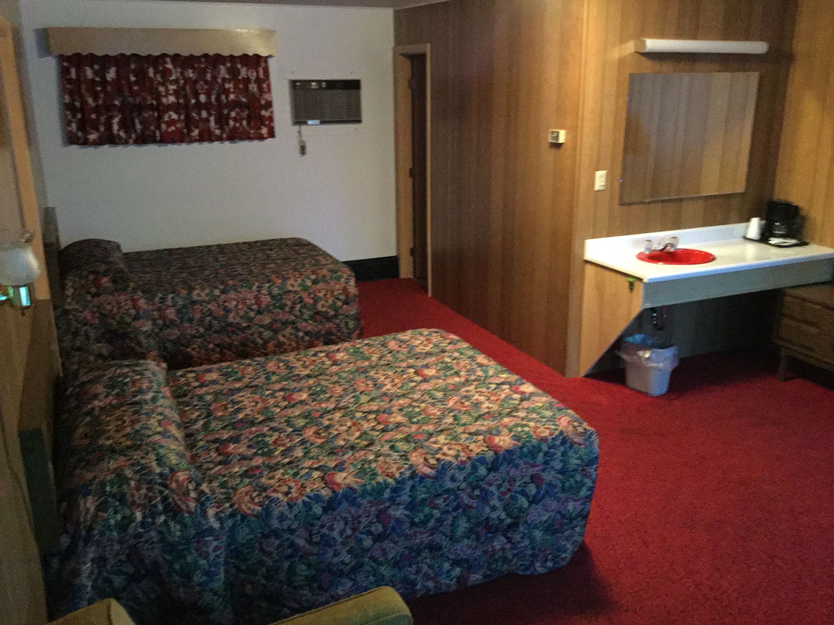 A room with two beds, a desk with a red sink, a mirror, and a trash bin.