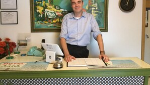 Un hombre de pie detrás de un mostrador de recepción, con un cuadro, diplomas enmarcados y un reloj en la pared.