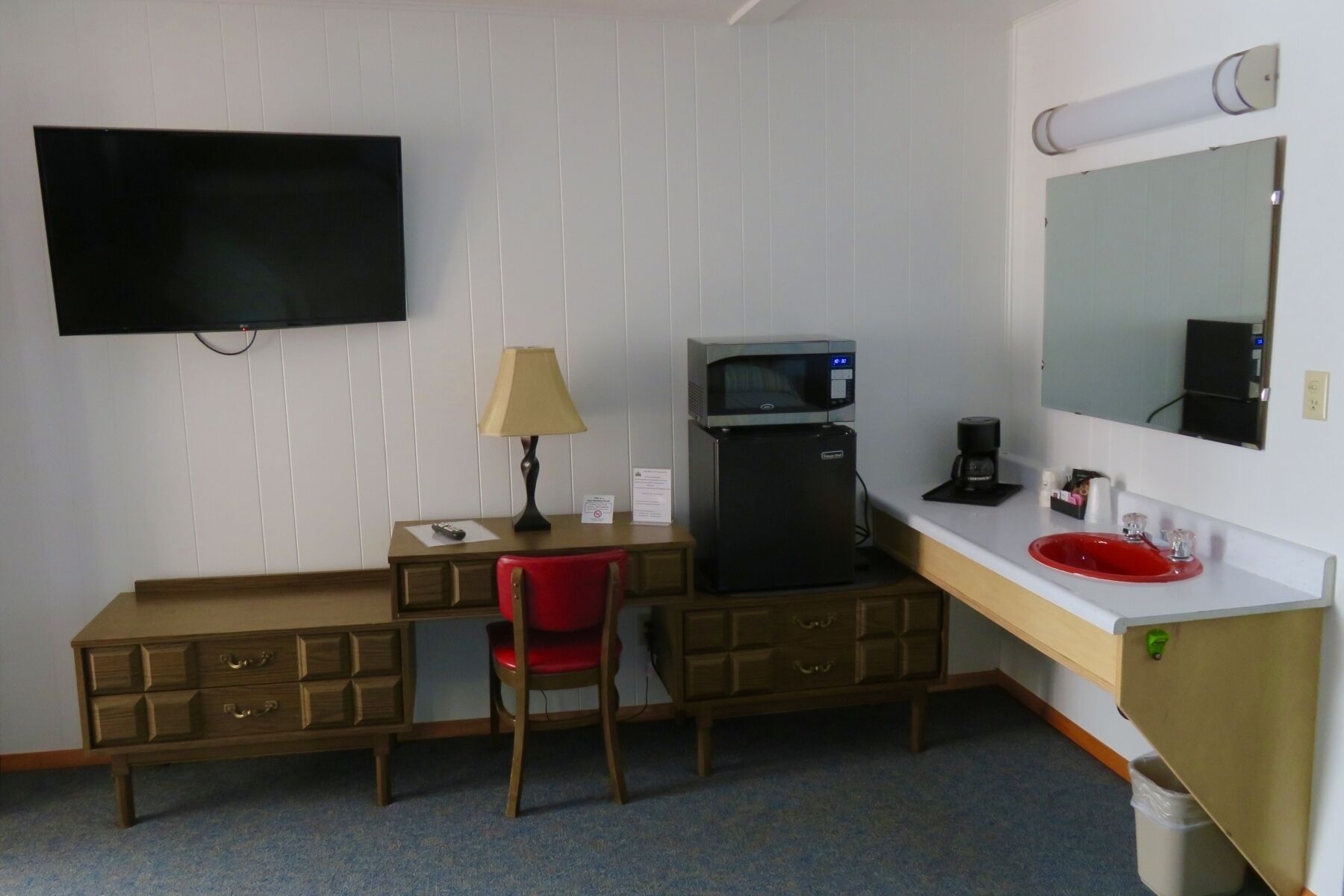A compact hotel room with a single bed, a desk with a chair, a microwave, a coffee maker, a TV, and a red sink.