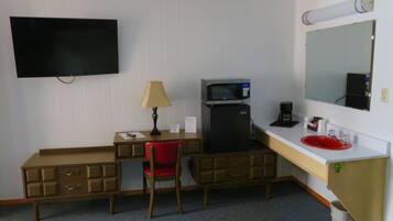 A compact hotel room with a single bed, a desk with a chair, a microwave, a coffee maker, a TV, and a red sink.