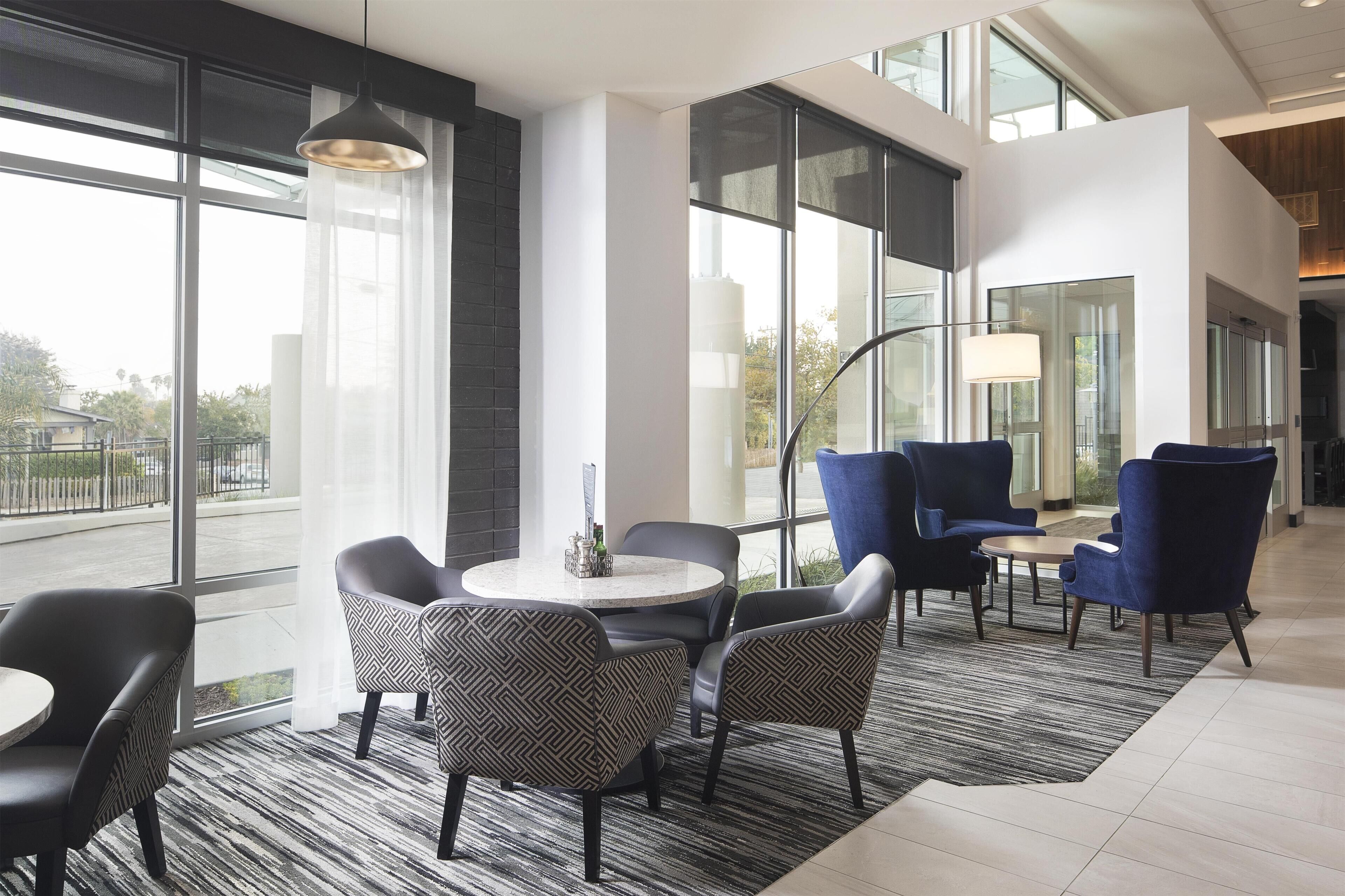 A modern hotel lobby with large windows, a round table with chairs, and a blue sofa area.