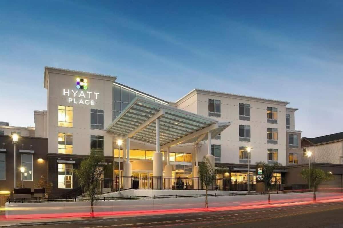 Un hotel de varios pisos con un toldo de cristal en la entrada, un restaurante visible y un estacionamiento.