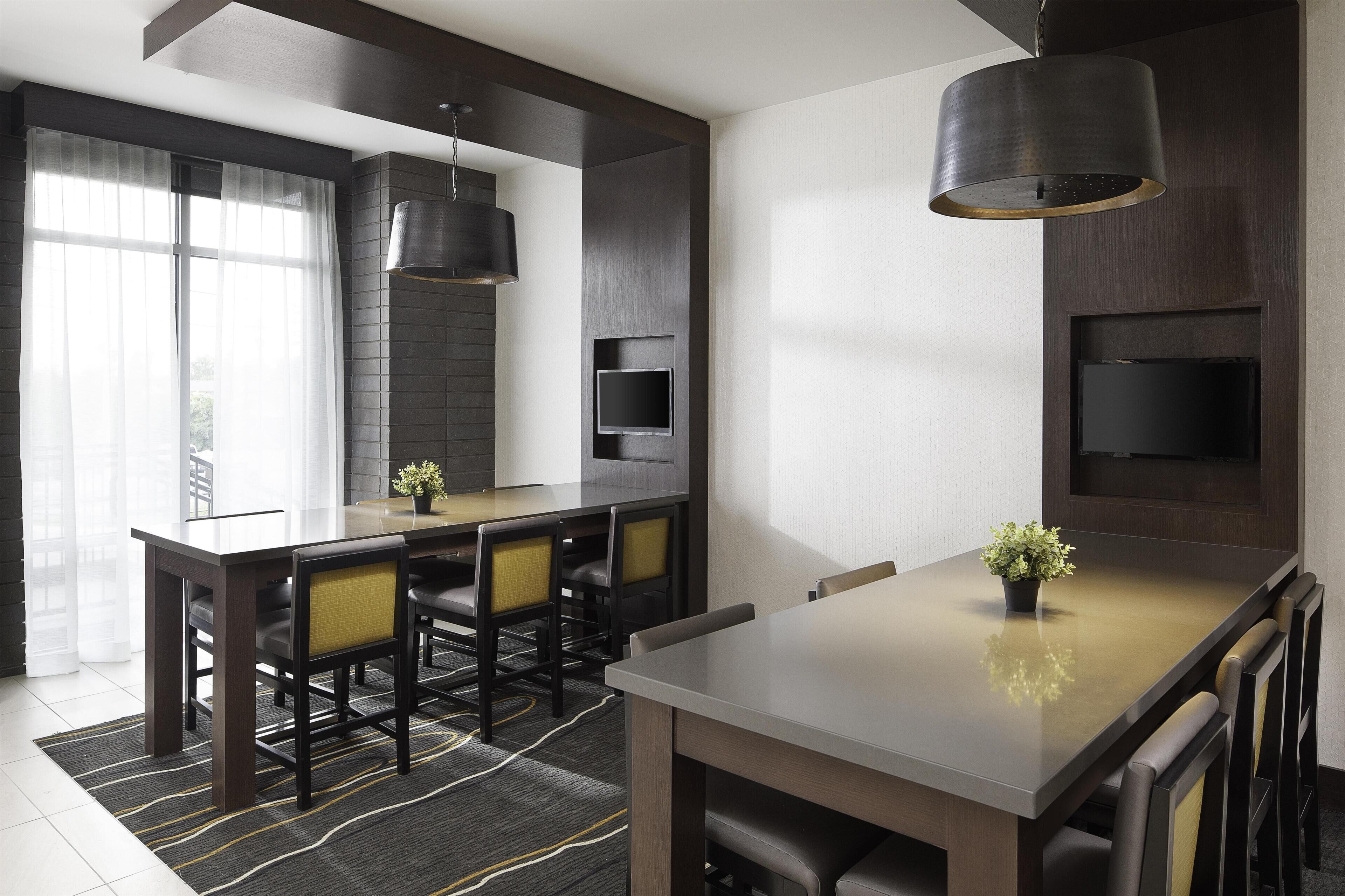 A modern dining area with a long table, chairs, a hanging light fixture, and a flat-screen TV mounted on the wall.