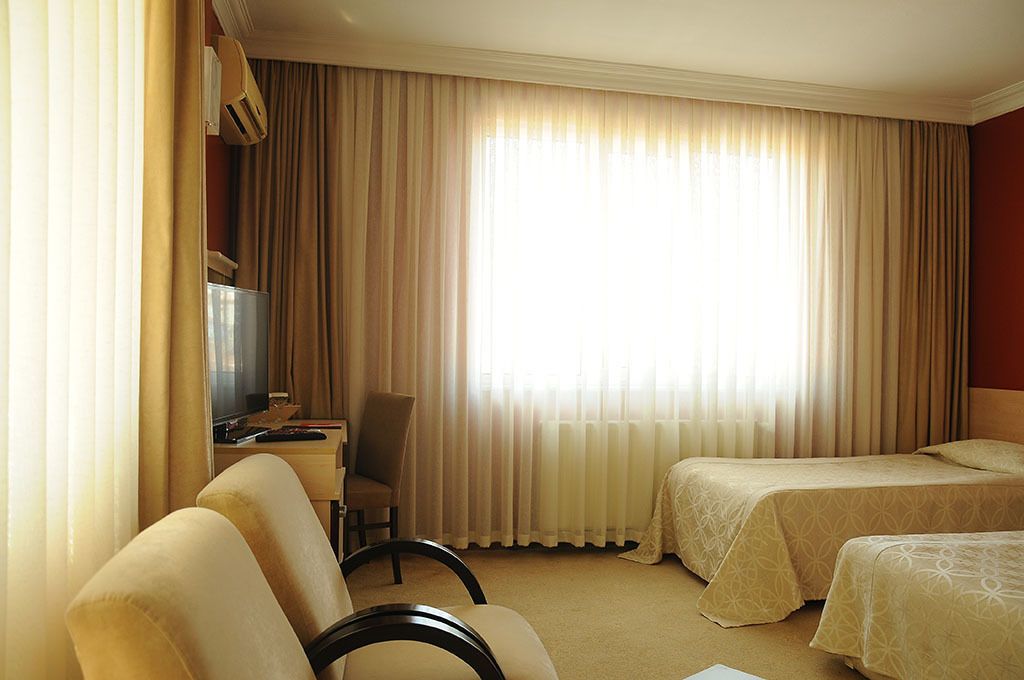 A hotel room with two beds, a desk with a chair, a television, and a large window with curtains.